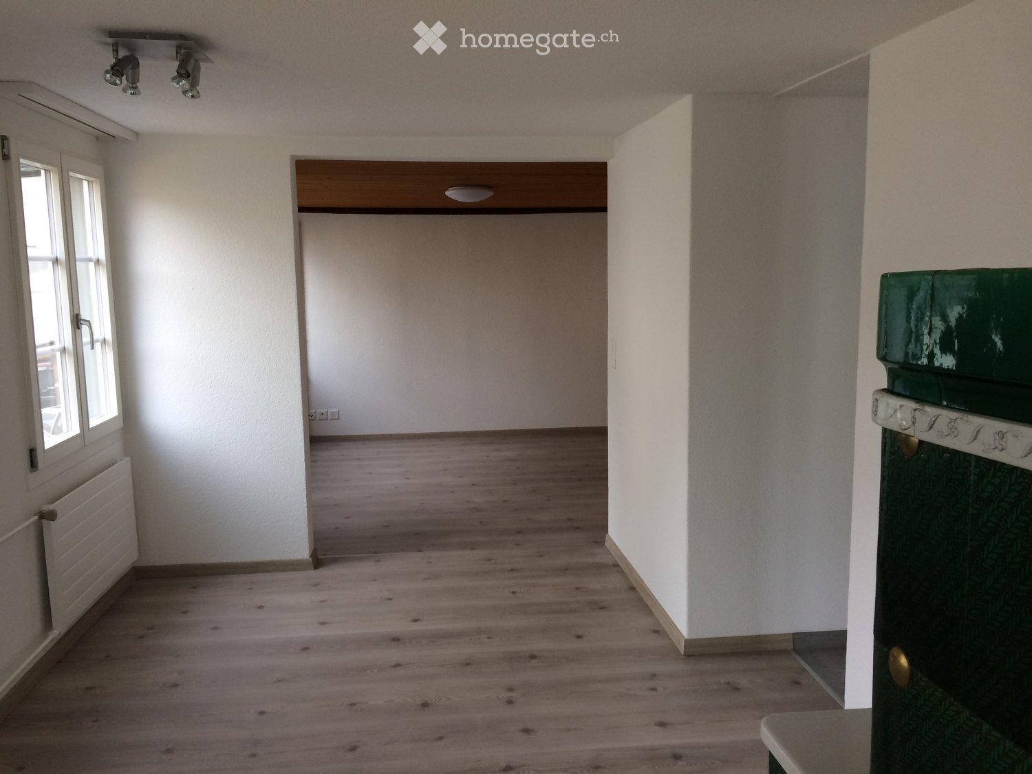 Empty room with wooden flooring, white walls, and a window on the left side. The room has a door leading to another space and a radiator below the window.