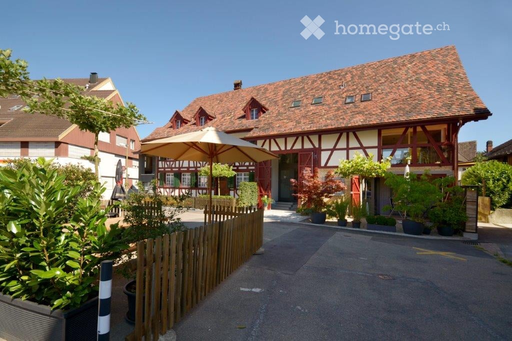 Two story brick house, red roof, large front yard, wooden fence, patio with umbrella, various potted plants