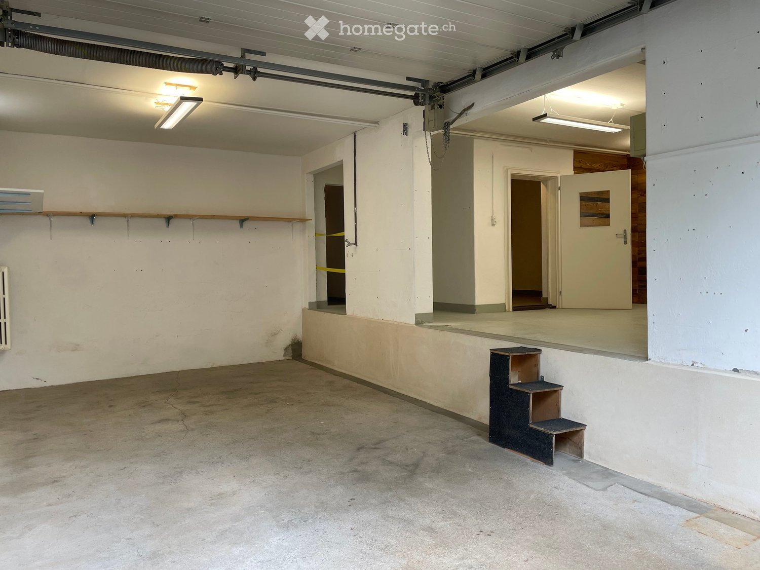 Empty garage with concrete floor, white walls, and two doors leading to other areas. Minimal lighting and a small step stool near the wall.