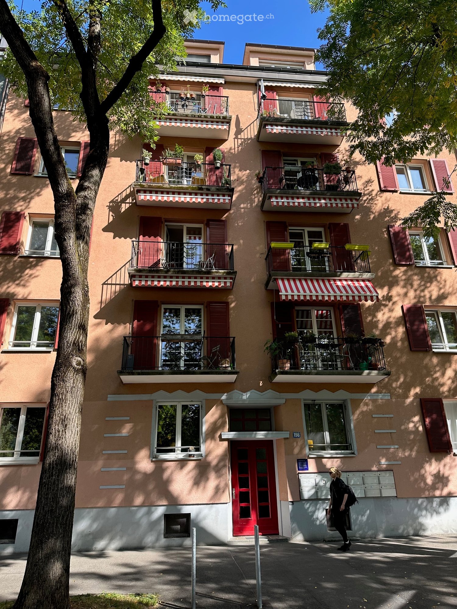Four-story residential building with multiple balconies, red shutters, and a central entrance. Balconies have outdoor furniture and potted plants.