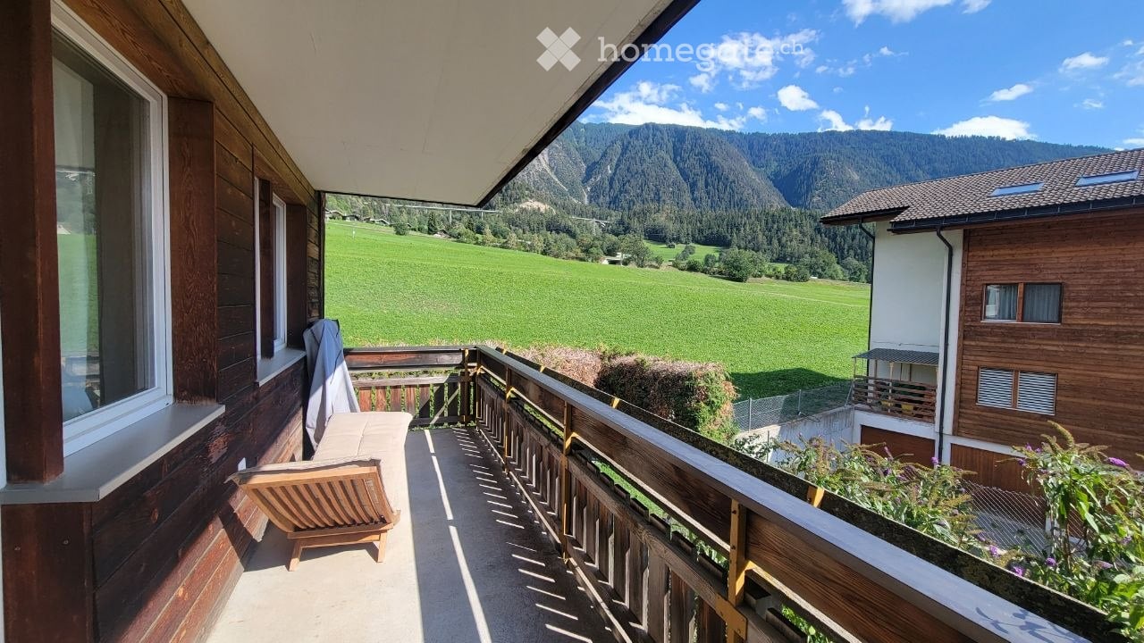 Balcony with wooden railing, overlooking a green valley and mountains, with a wooden chair and small table.