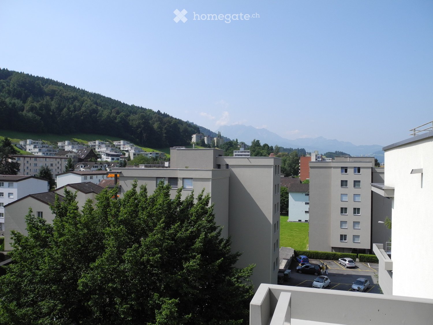 View of a residential area with multiple apartment buildings, a parking lot, and surrounding greenery with mountains in the background.