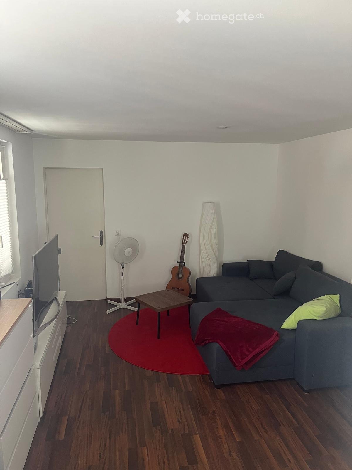 Modern living room with wooden flooring, a grey sectional sofa with cushions, a small wooden coffee table, a red circular rug, a standing fan, a guitar, and a television on a wooden stand.