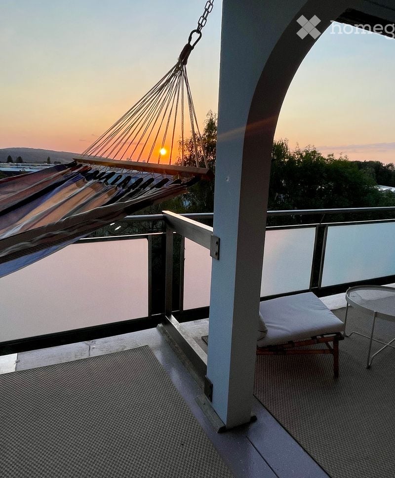 Balcony with a hammock, lounge chair, and small table, overlooking a scenic sunset view.