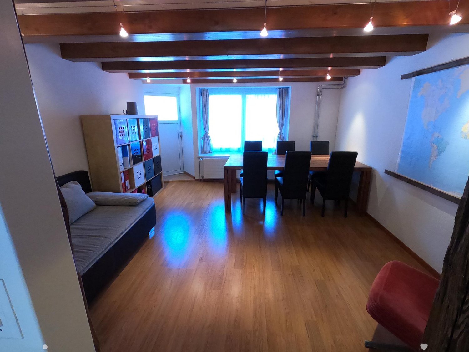 A multi-functional room with a sofa, dining table, chairs, and a storage unit. The room features wooden flooring and large windows with curtains.