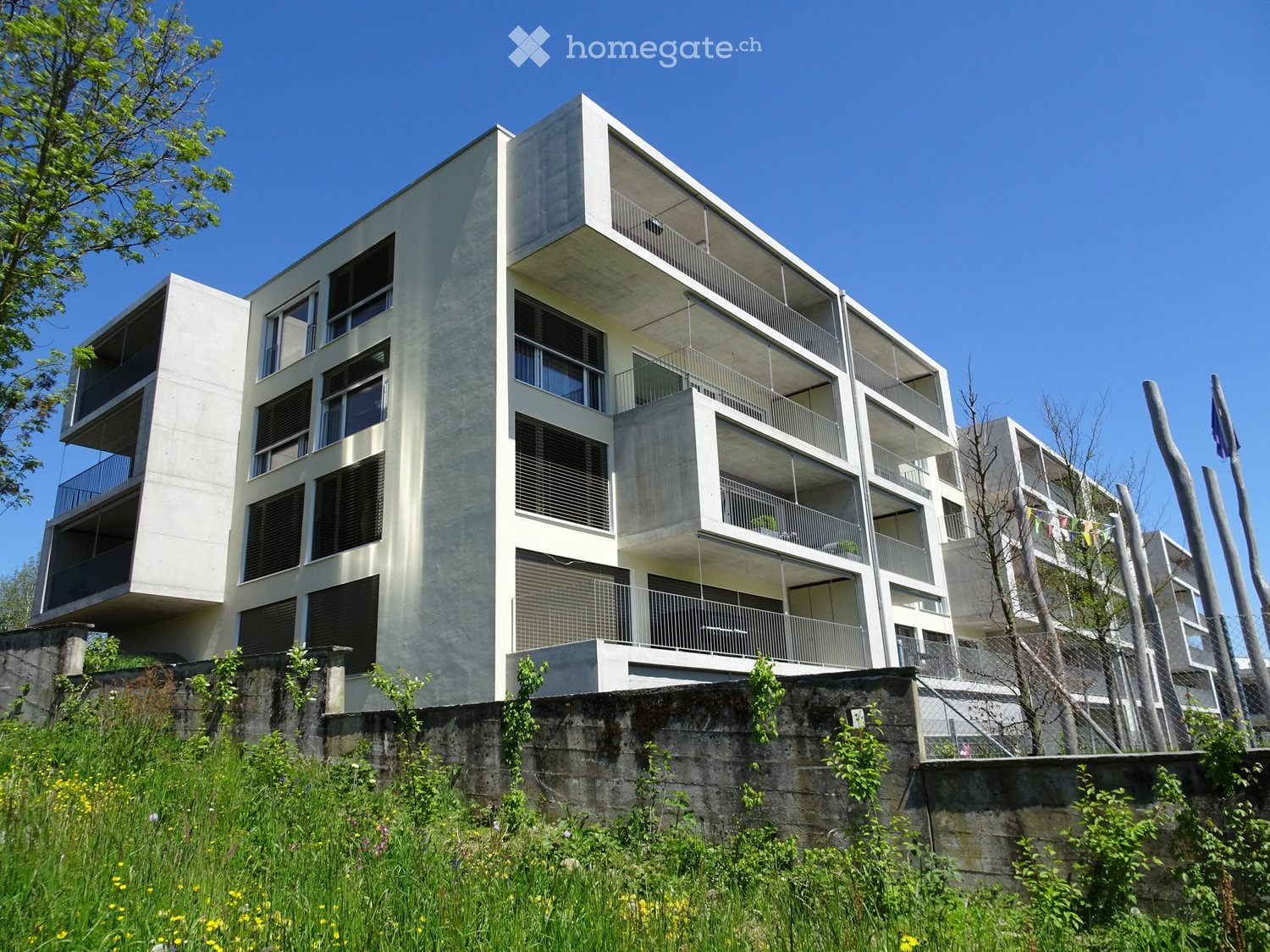 Modern multi-story residential building with balconies and large windows, surrounded by greenery and a stone retaining wall.