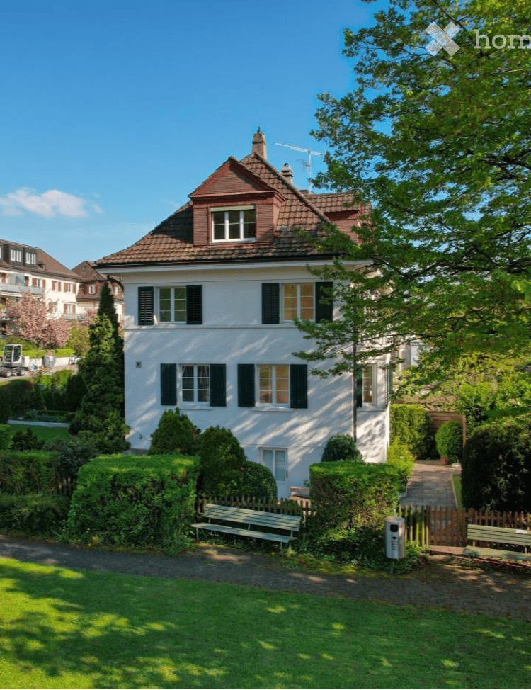 A two-story white house with a red-tiled roof, multiple windows with shutters, and a well-maintained garden with trimmed hedges, a bench, and a wooden fence.