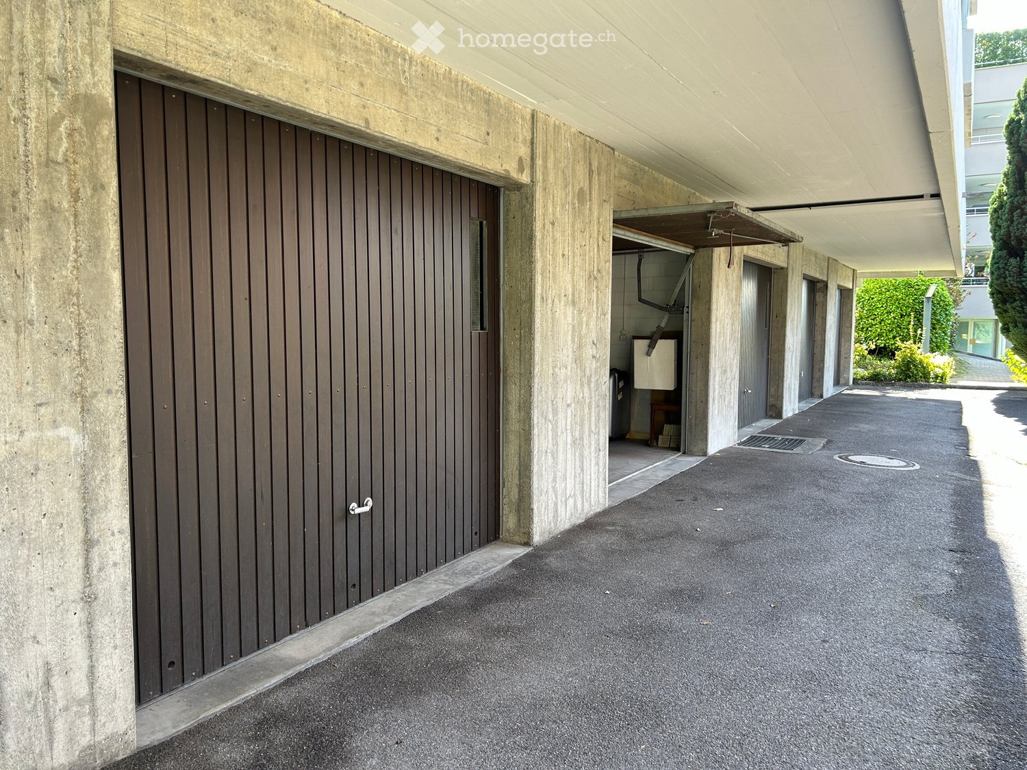 Modern building entrance with a covered concrete walkway, a wooden garage door, and a small open garage area with a white appliance visible inside.