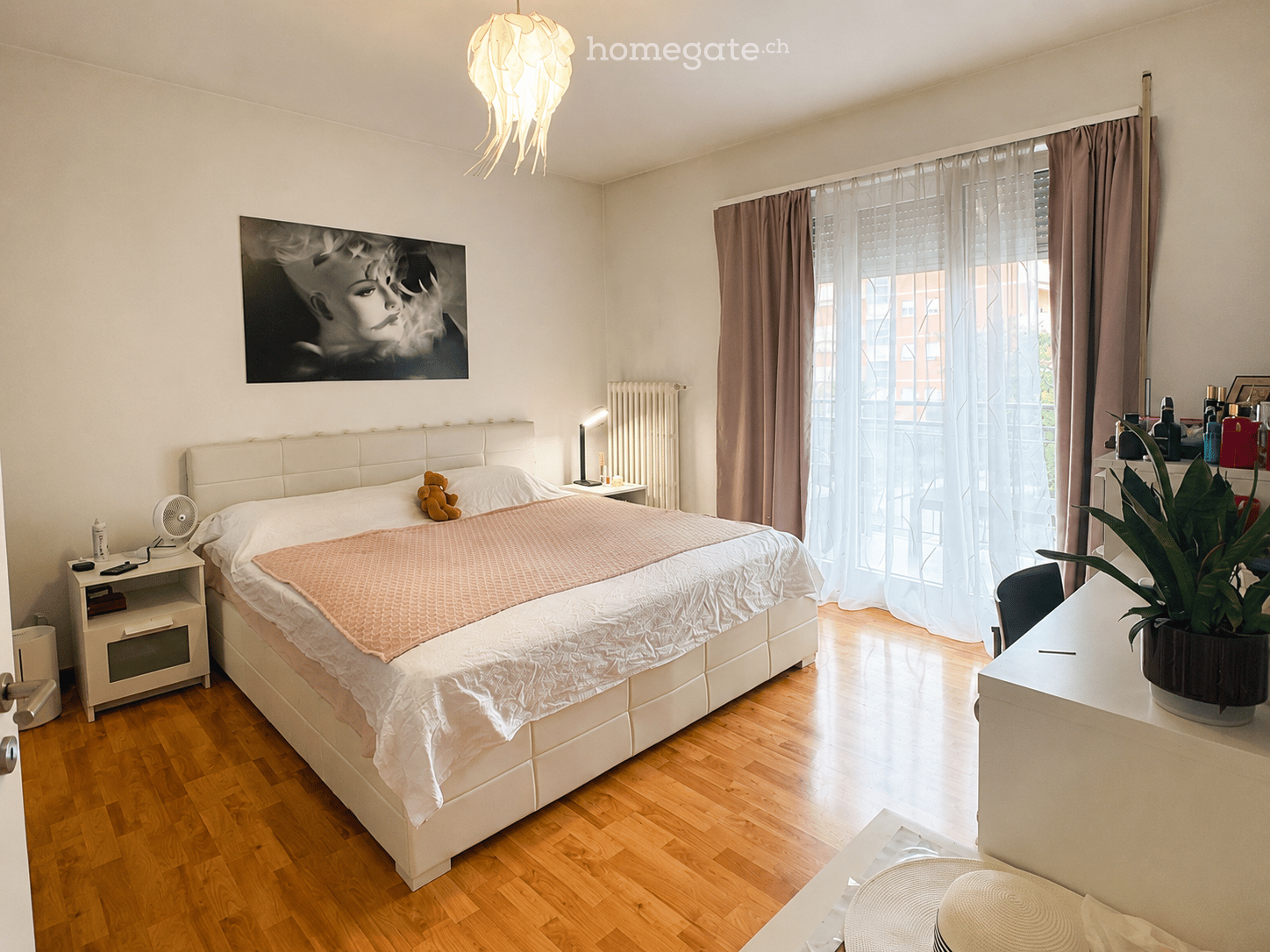 Modern bedroom with a large bed, nightstand, fan, desk, potted plant, and artwork on the wall.
