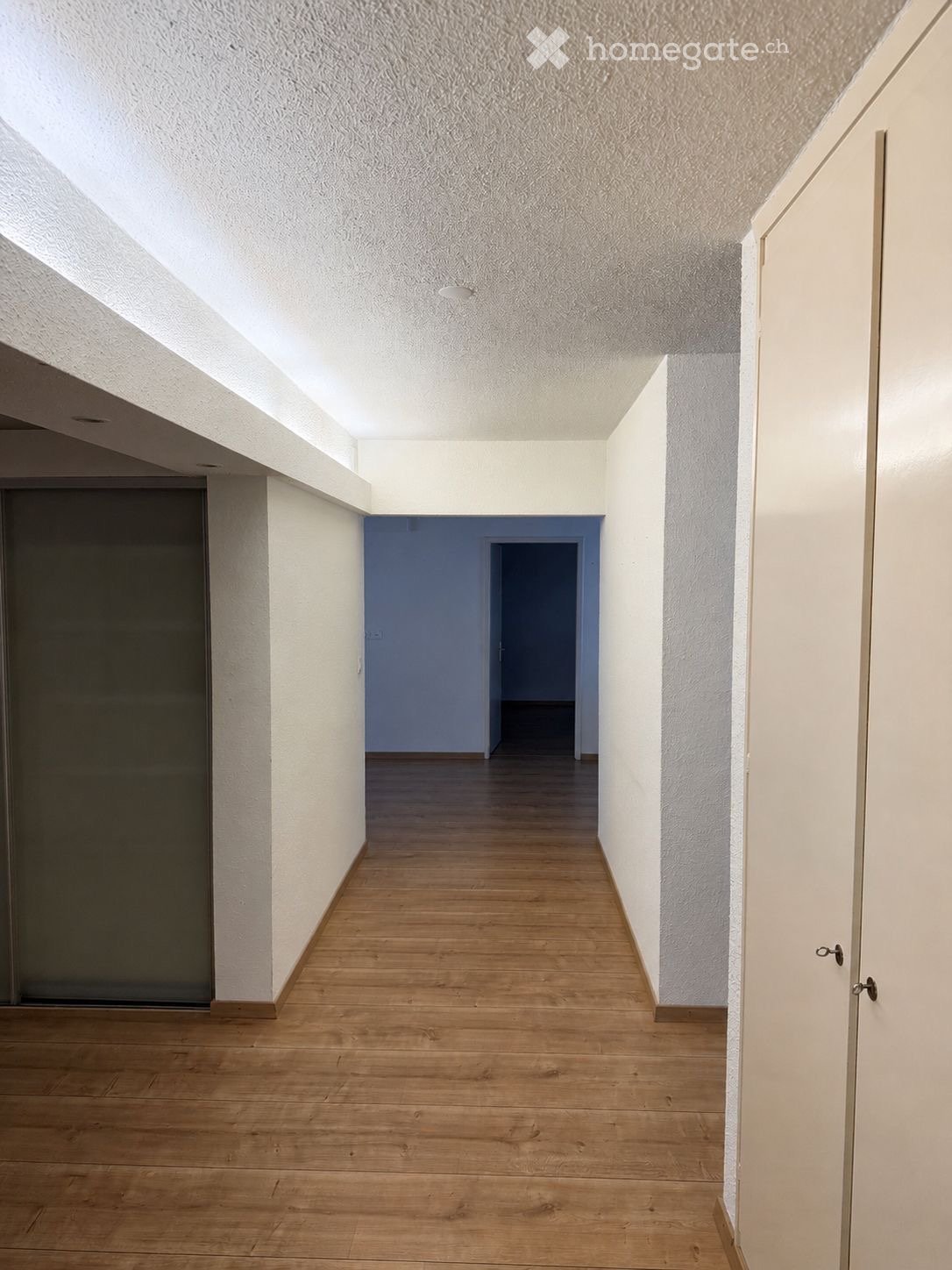 Empty hallway with wooden flooring, white walls, and a blue accent wall leading to another room.