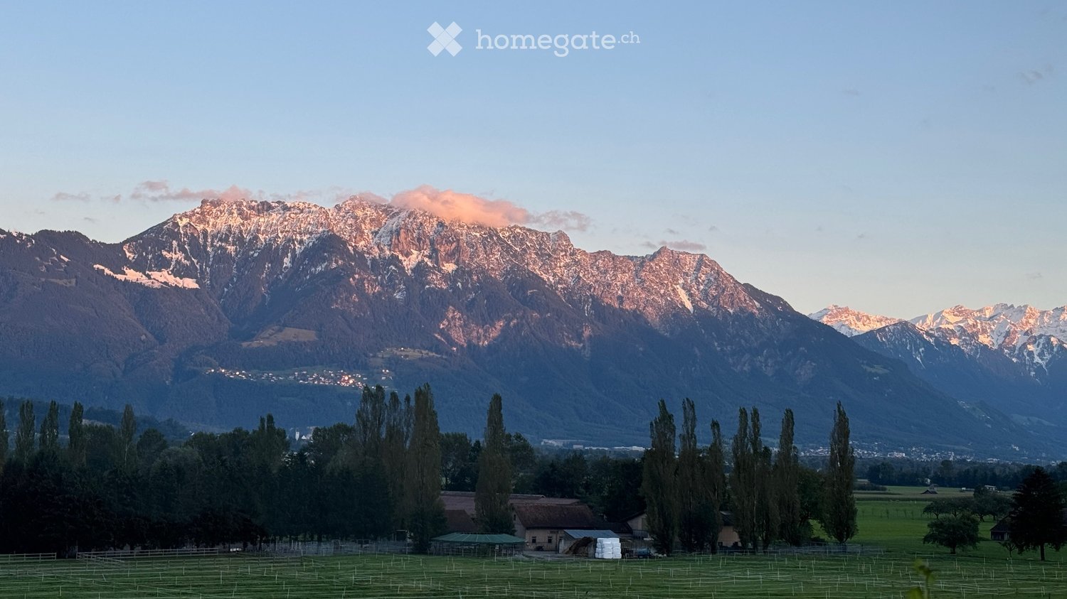 A serene outdoor scene featuring a lush green field in the foreground, bordered by tall trees, with a small house and barn visible. In the background, majestic snow-capped mountains rise under a clear blue sky.