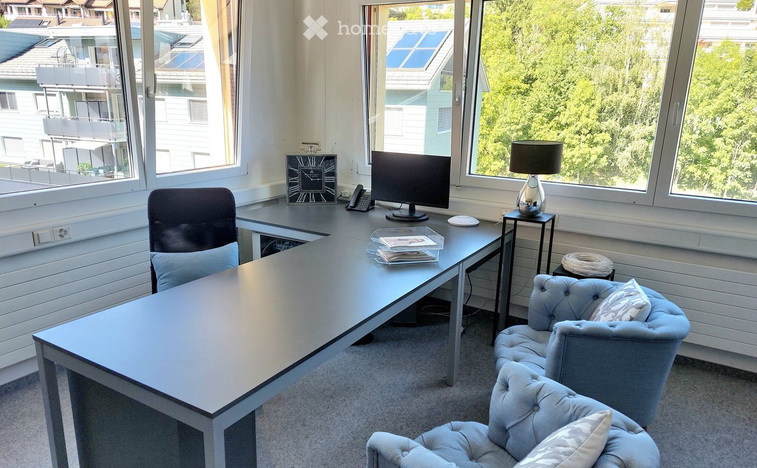 Office space with a large desk, computer screen, office chair, two armchairs, a lamp, a clock, and a telephone. Large windows offer a view of greenery and neighboring buildings.