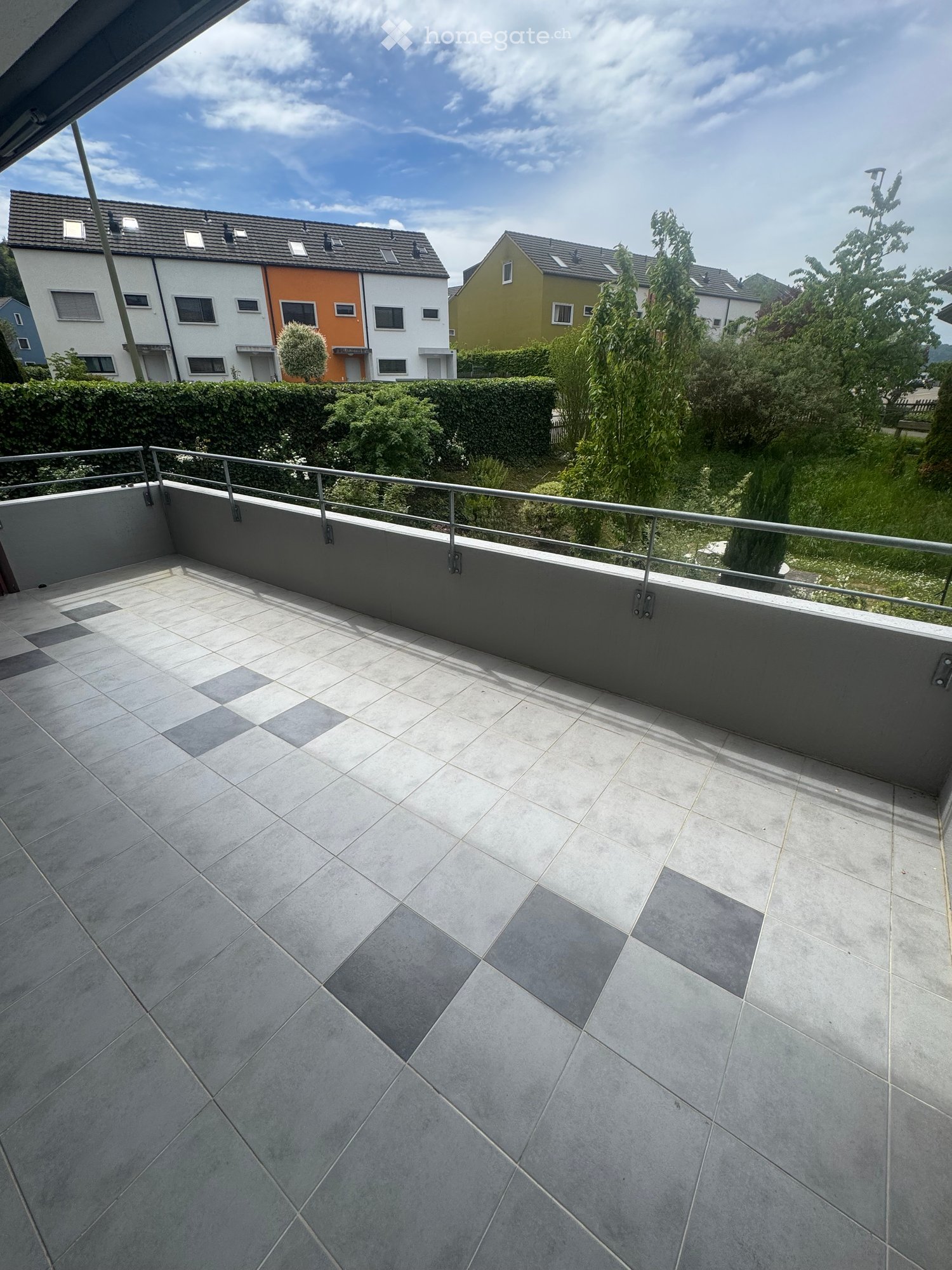 Modern balcony with grey tiled flooring, metal railing, and a view of neighboring houses and greenery.