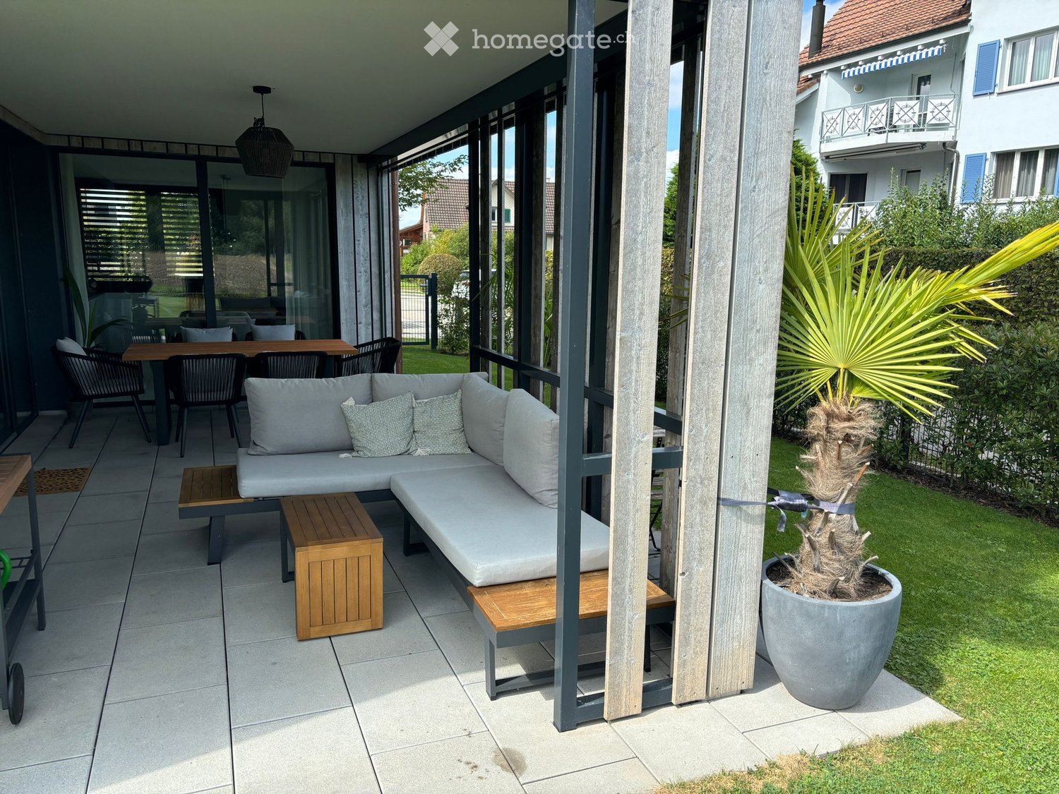 Balcony with a grey sectional sofa, wooden stools, a dining table with chairs, and a potted plant.