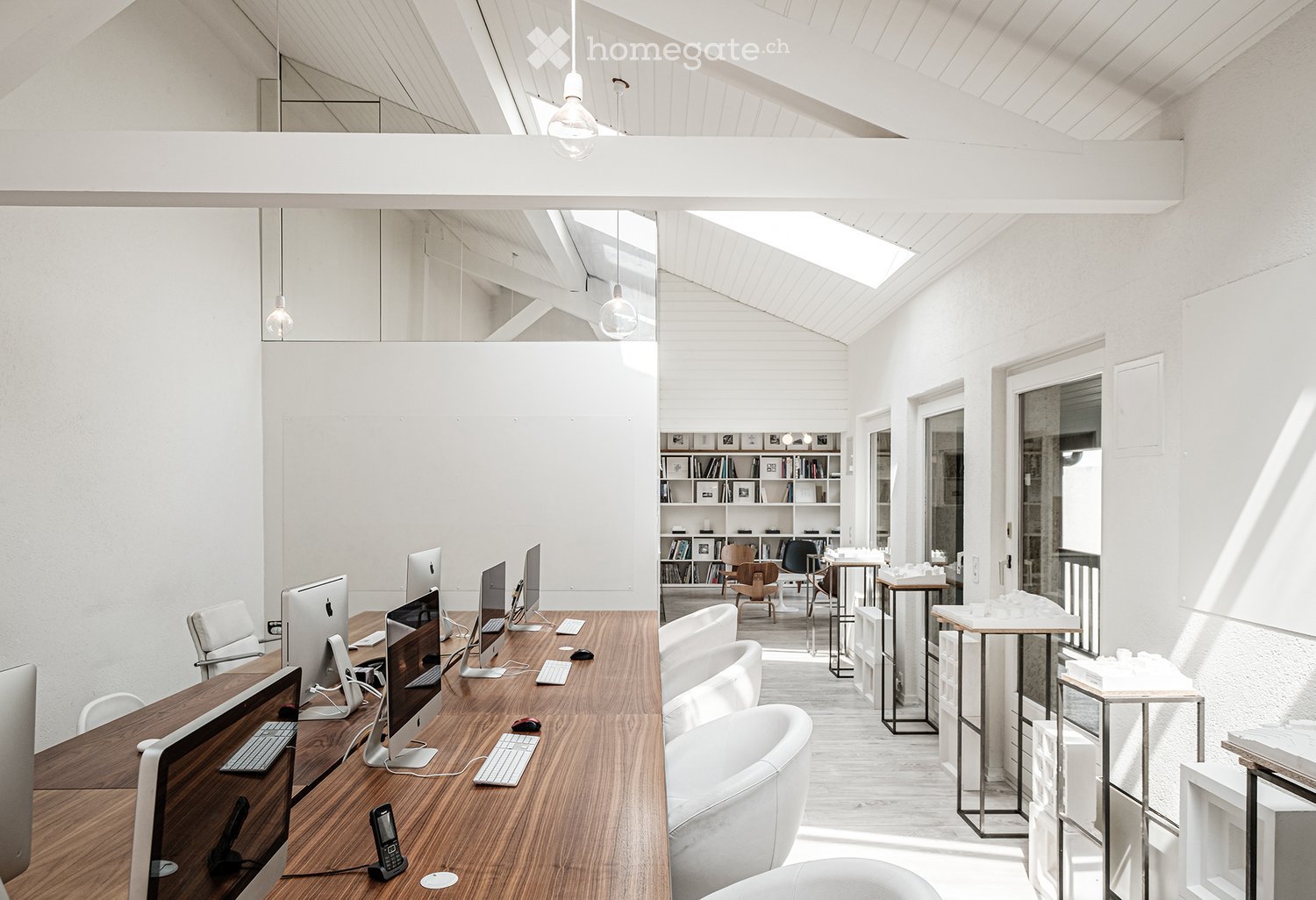 Bright, open-plan office space with wooden desks, multiple computer screens, keyboards, mice, and white chairs. Shelves with books and decorative items are visible in the background.