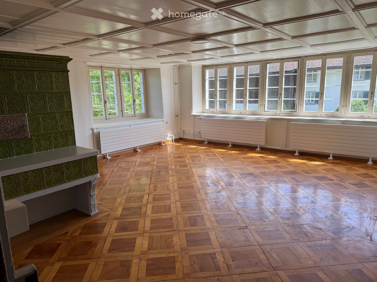 Unfurnished Room. Empty room with wooden parquet flooring, large windows, radiators, and a decorative green fireplace.