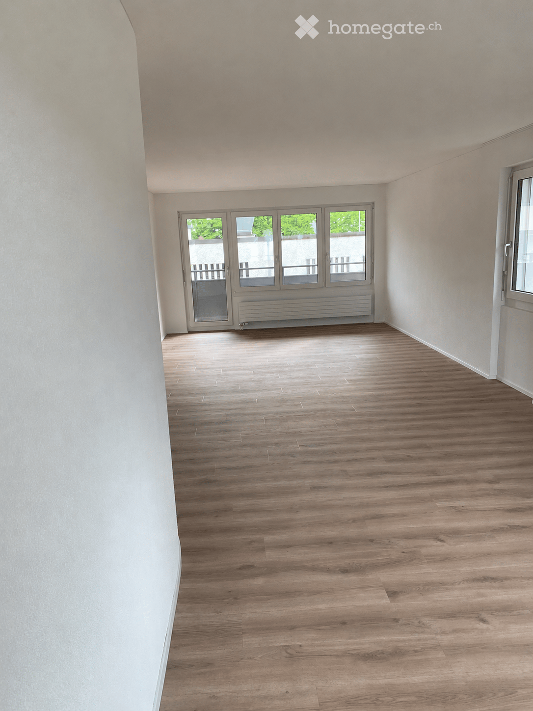 Empty room with light wooden flooring, white walls, and large windows with a view of greenery. No furniture or equipment is present.