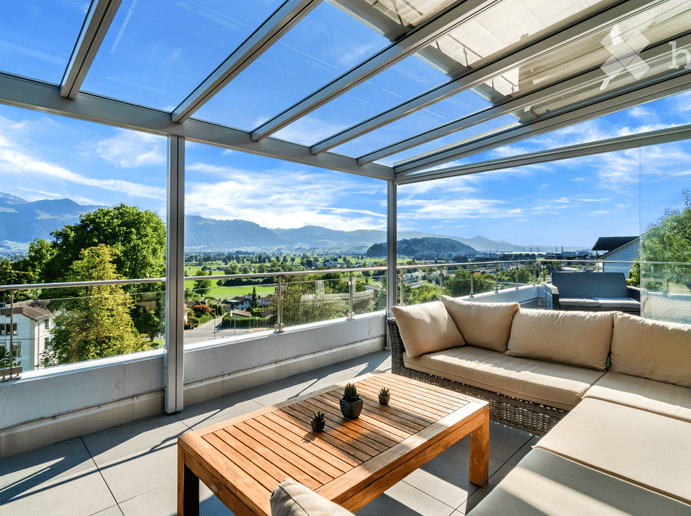 A modern glass-enclosed balcony with a panoramic mountain view, featuring a wooden coffee table, a cream-colored sofa, and small potted plants.