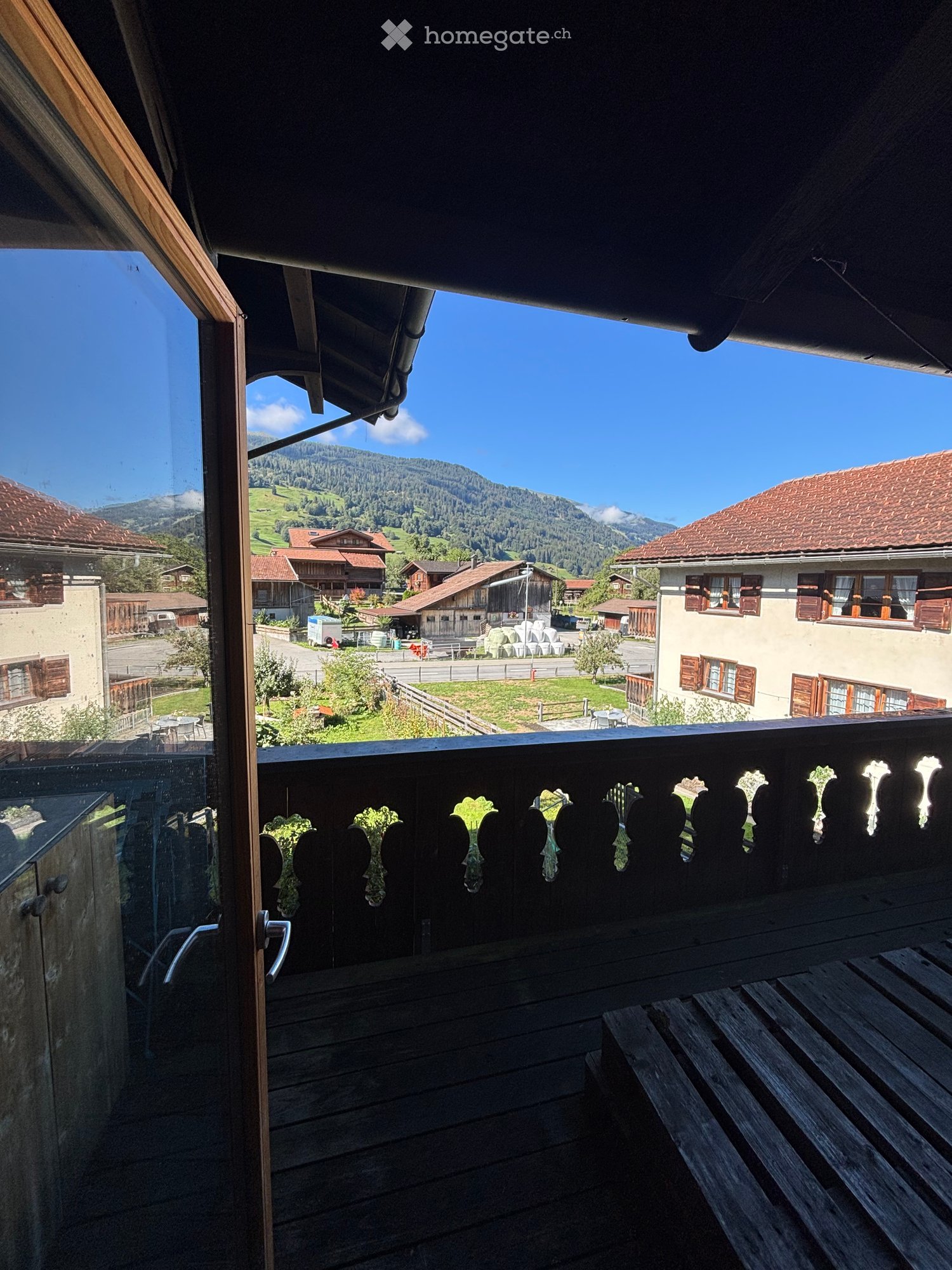 Balcony with wooden decking, ornate wooden railing, and scenic mountain view of a village with traditional houses and green fields.
