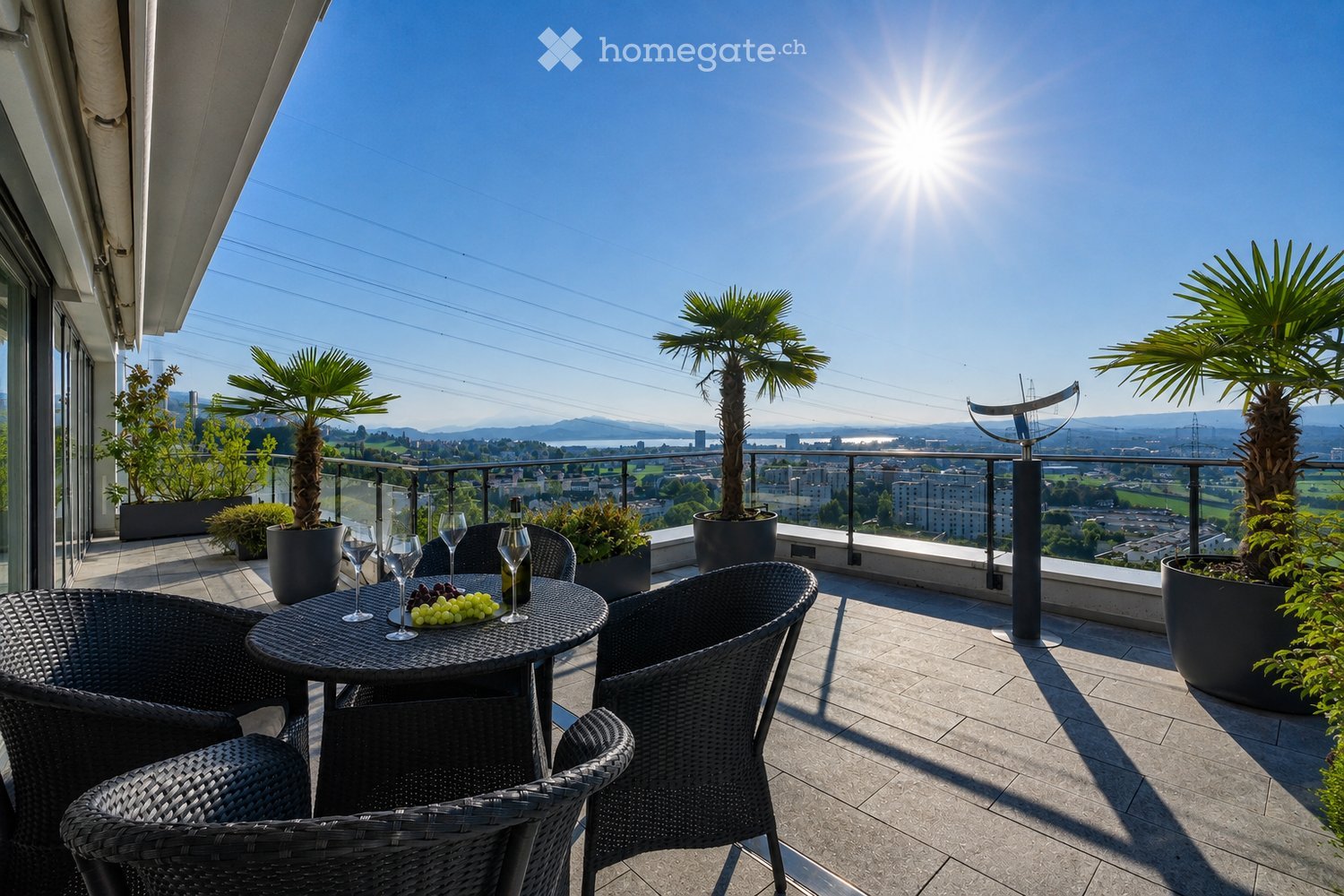 Balcony with a round table, four wicker chairs, wine glasses, a bottle, and a bowl of grapes, surrounded by potted plants and offering a scenic city view.