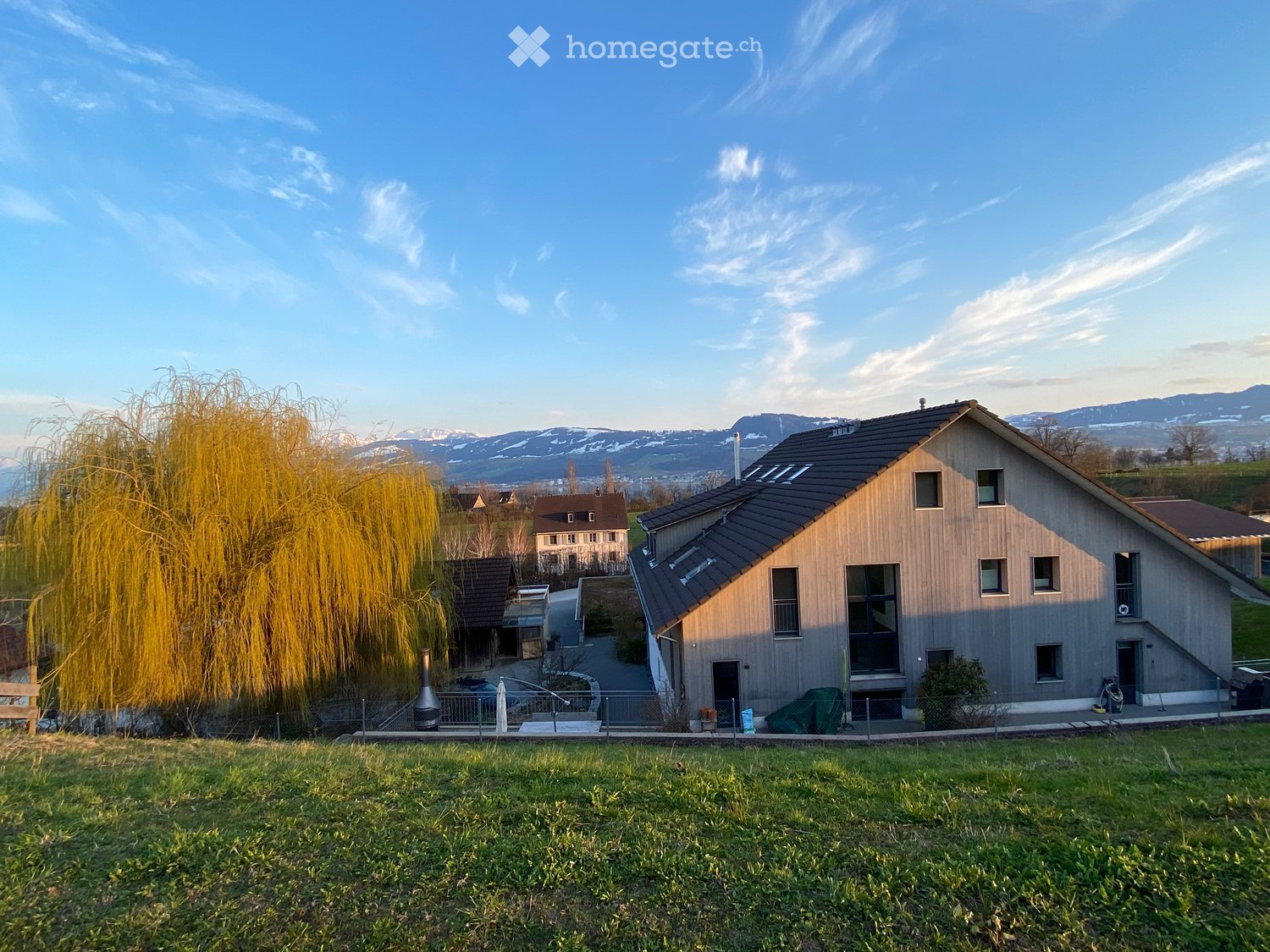 A modern two-story house with a wooden facade, multiple windows, and a dark roof, situated on a grassy slope with a scenic mountain view in the background.