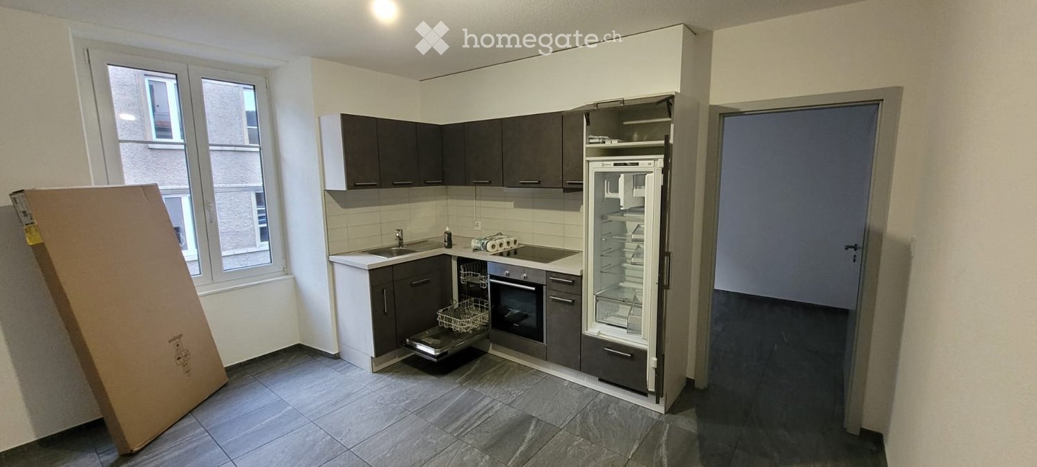 Modern kitchen with grey cabinets, white countertops, stainless steel appliances, and a large window. A cardboard box is visible on the left side.