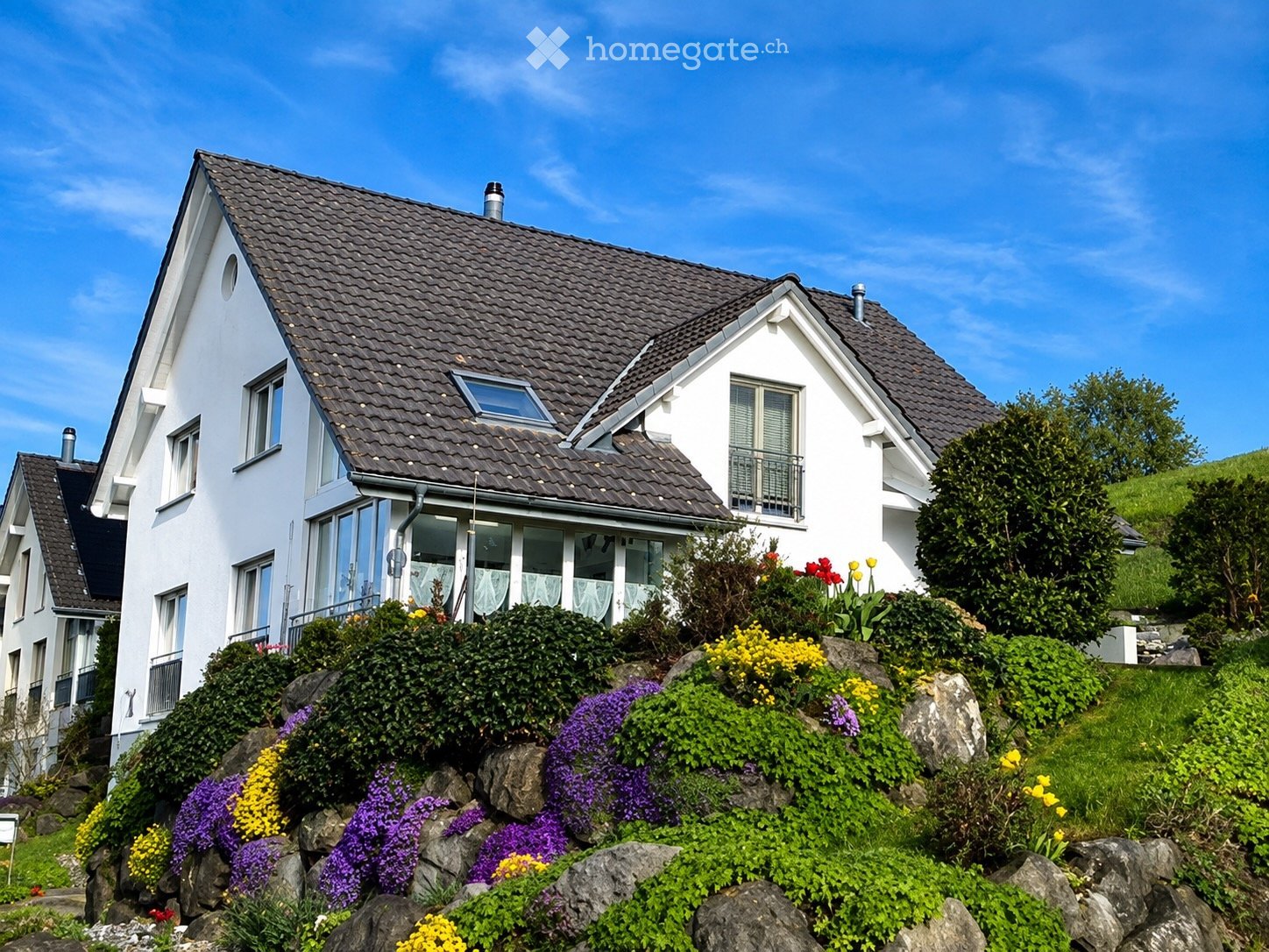 A modern two-story white house with a dark shingled roof, multiple windows, and a balcony. The house is surrounded by a well-maintained garden with colorful flowers and shrubs.