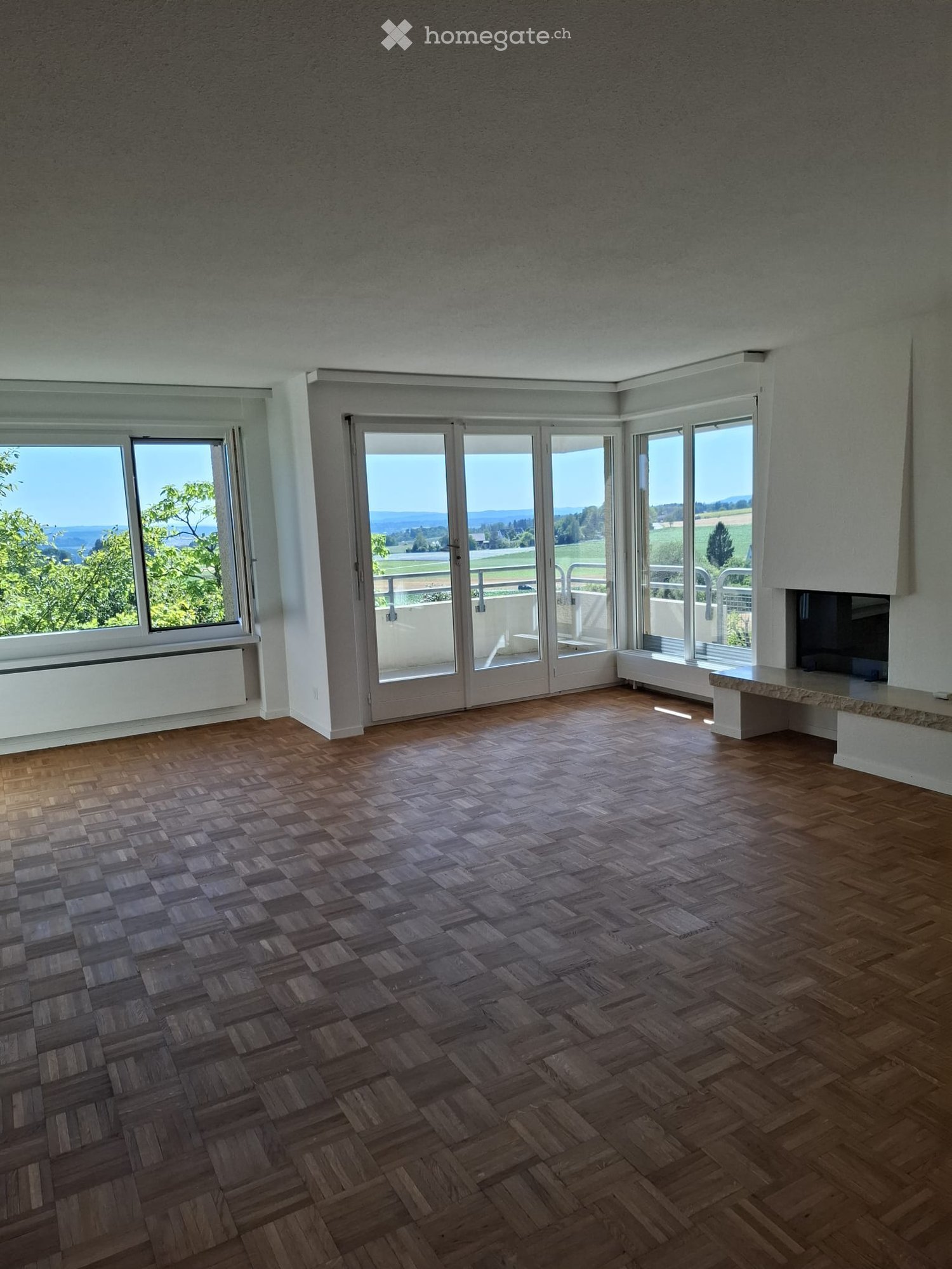 Empty room with wooden herringbone flooring, large windows and sliding glass doors leading to a balcony, offering scenic views of greenery and fields.