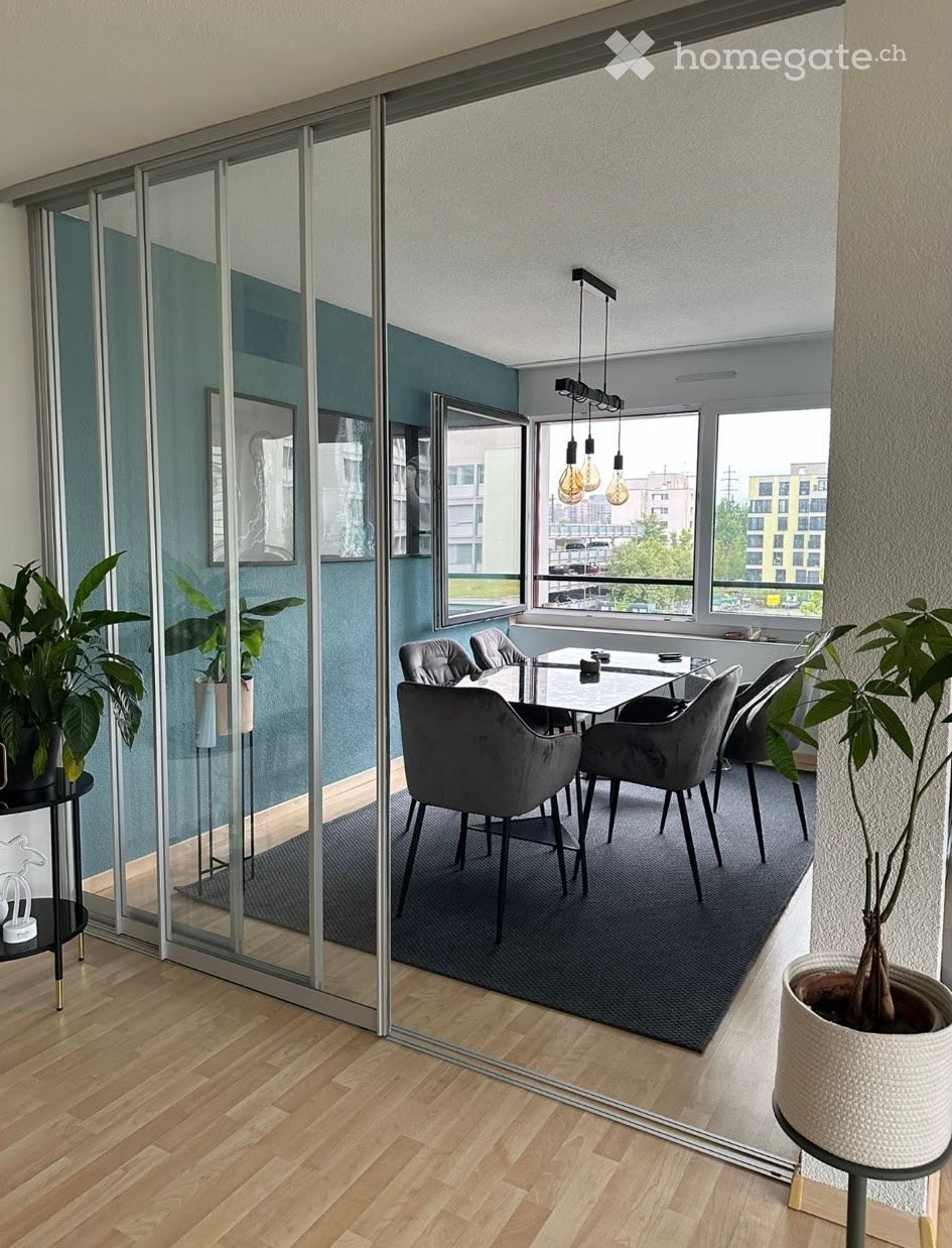 Modern dining area with a sleek glass table, grey upholstered chairs, large windows, and potted plants.