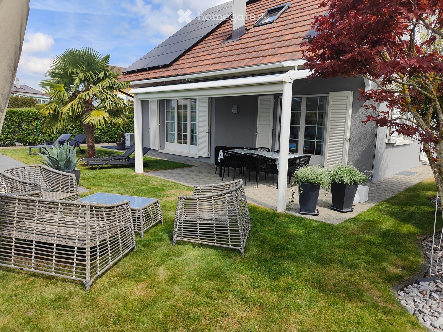 Well-maintained garden with a wicker sofa, wicker chairs, coffee table, dining table with chairs, sun loungers, potted plants, and a modern house with large windows and solar panels on the roof.