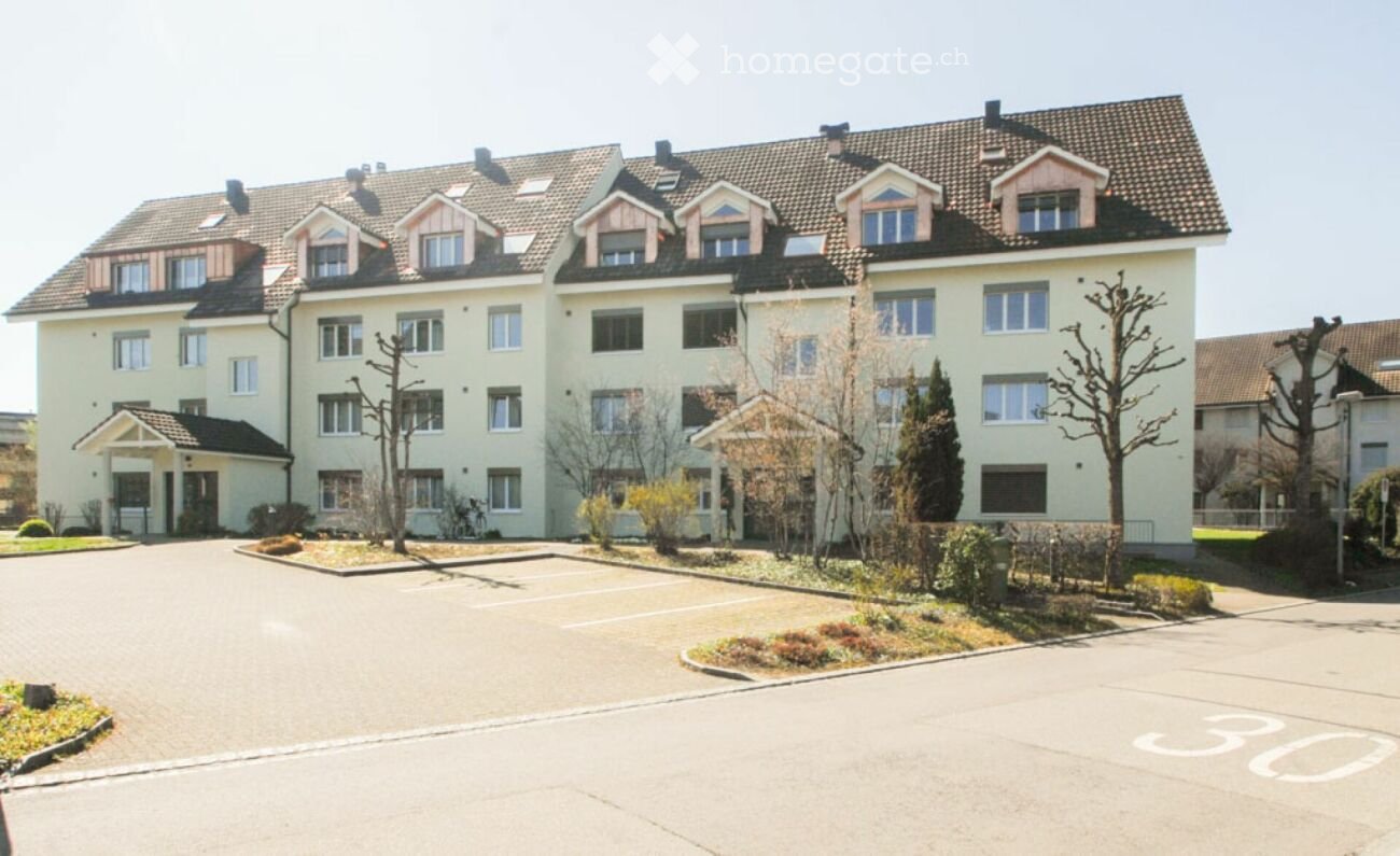 Multi-story residential building with light yellow facade, multiple windows, and a parking area in front.