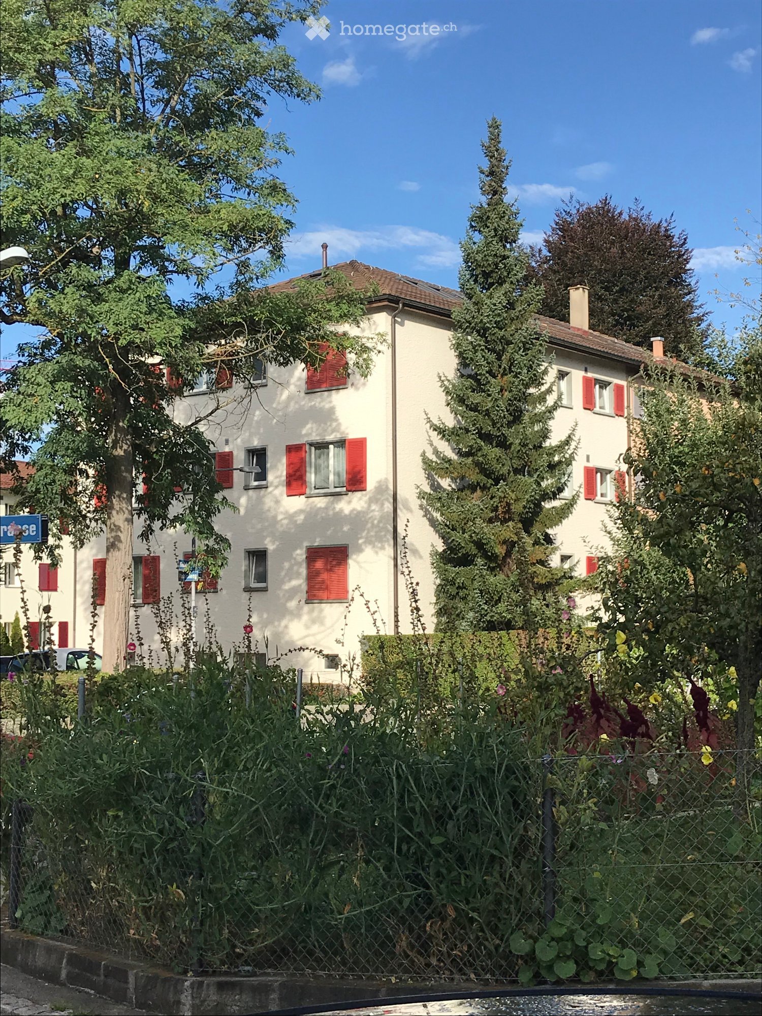 Two-story house with beige walls, red shutters, and multiple windows, surrounded by trees and greenery.