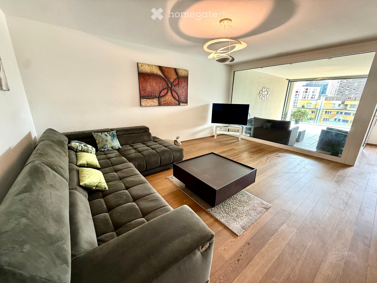 Modern living room with a large sectional sofa, coffee table, TV, and artwork on the wall. The room features wooden flooring and large windows with a view of the city.