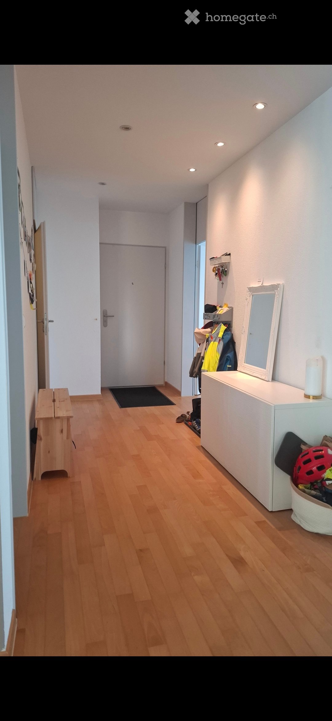 A well-lit hallway with wooden flooring, white walls, and recessed ceiling lights. Features include a coat rack with jackets, a small bench, a mirror on a console table, and a shoe rack with shoes and a helmet.