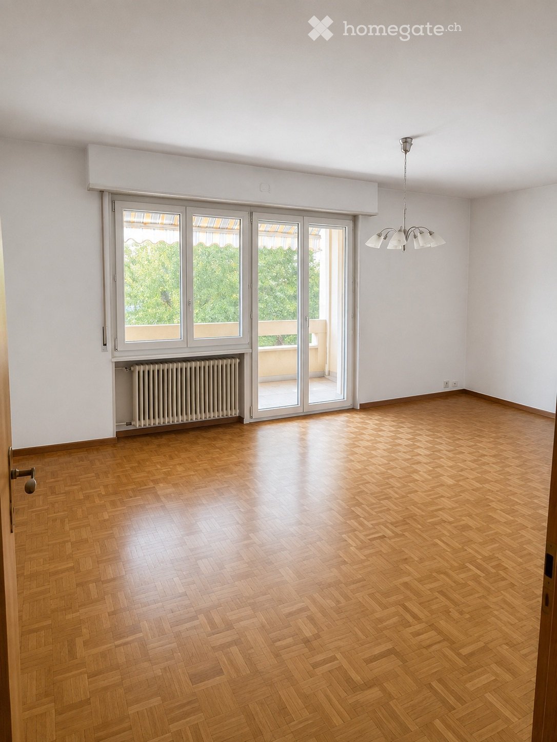 Empty room with wooden herringbone flooring, white walls, large sliding glass doors leading to a balcony, radiator, and a ceiling light fixture.