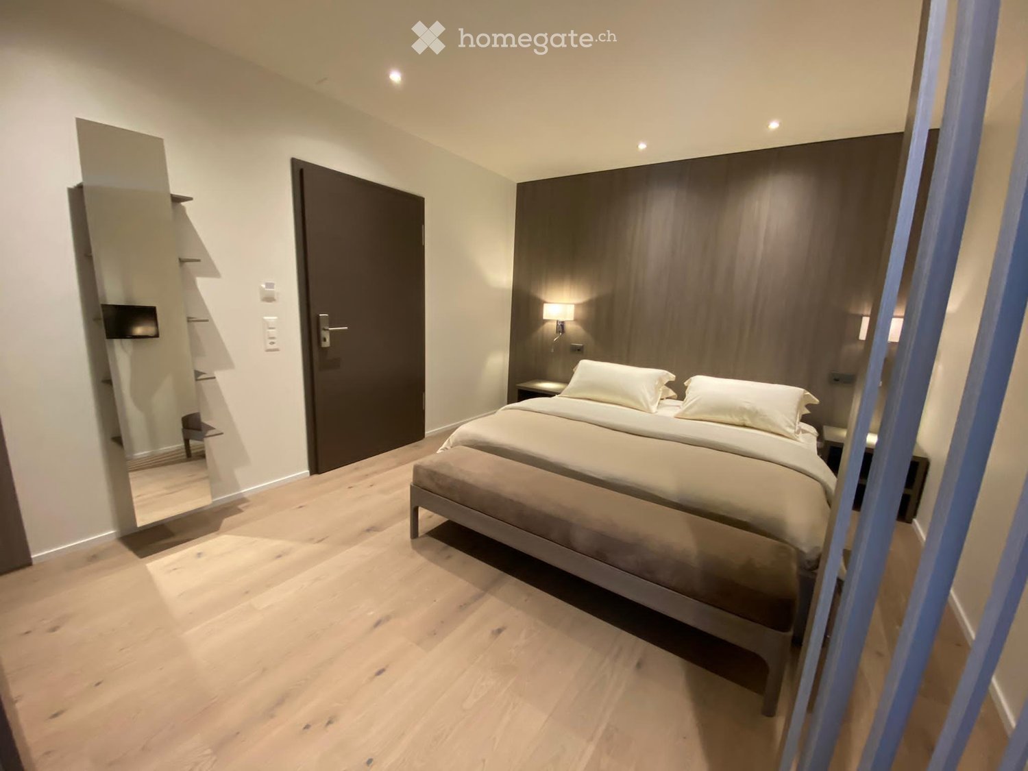 Wooden floor, double bed, 2 nightstands with lamps, wall-mounted mirror, wall-mounted TV, white walls, brown door