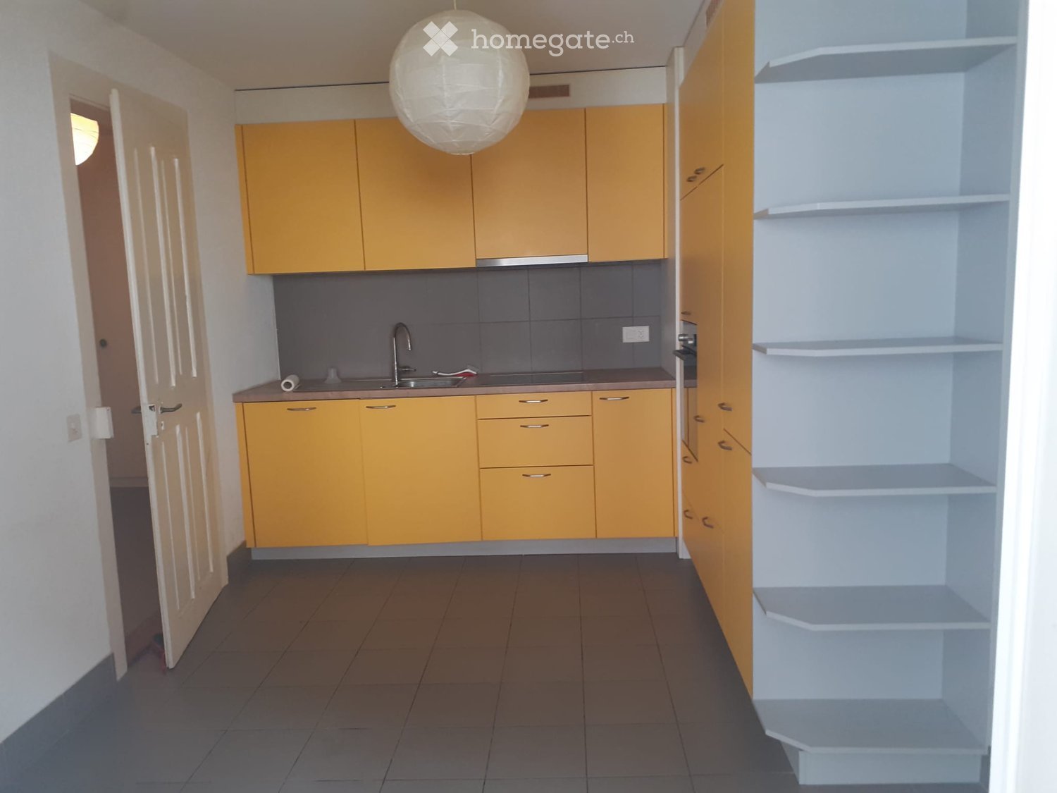 Modern kitchen with yellow cabinets, white shelves, and a sink with a faucet. The floor is tiled, and the space is well-lit with a hanging light fixture.