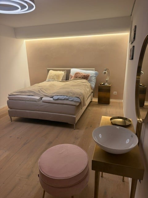 Modern bedroom with a large bed, nightstand with lamp, pink stool, and a small table with a bowl on it.