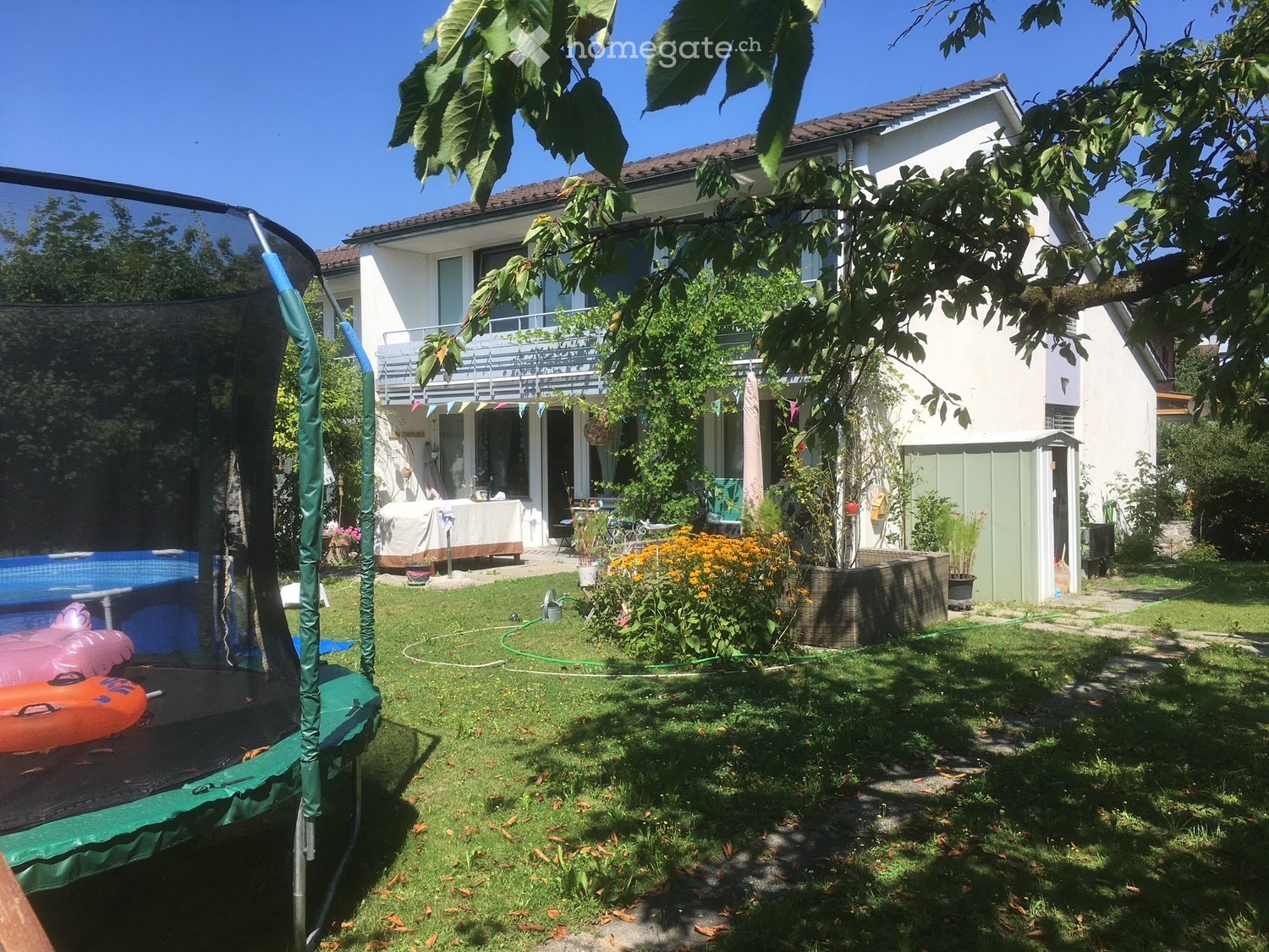 Lush garden with a trampoline, swimming pool, covered patio, and colorful flowers, surrounded by a two-story house with a balcony.