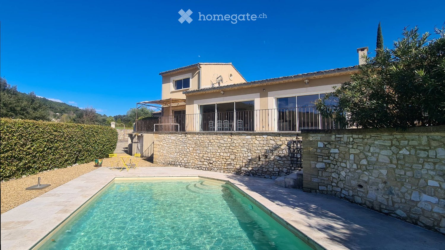 A modern house with a swimming pool in the foreground, surrounded by a stone wall and greenery. The house features large windows and a balcony with outdoor furniture.