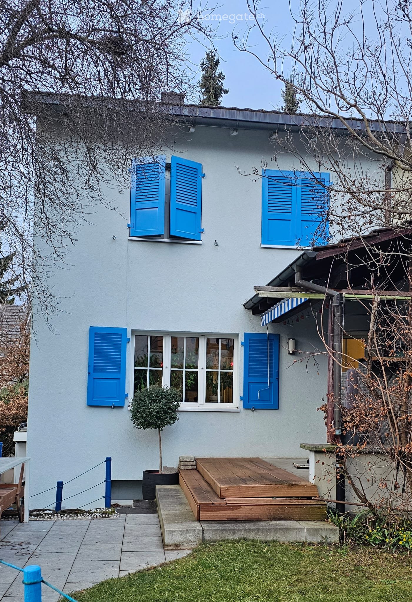 Two-story house with light blue shutters, a small wooden deck, and a small tree in a pot.