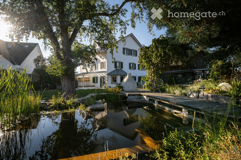 A serene garden with a pond reflecting the surrounding trees and house, featuring lush greenery, stepping stones, and a modern white sculpture.