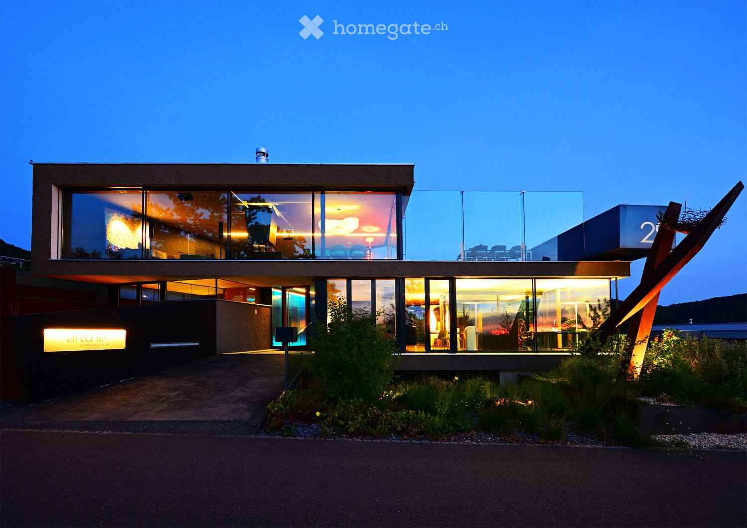 Modern two-story building with large glass windows, illuminated interior, and a landscaped garden area.