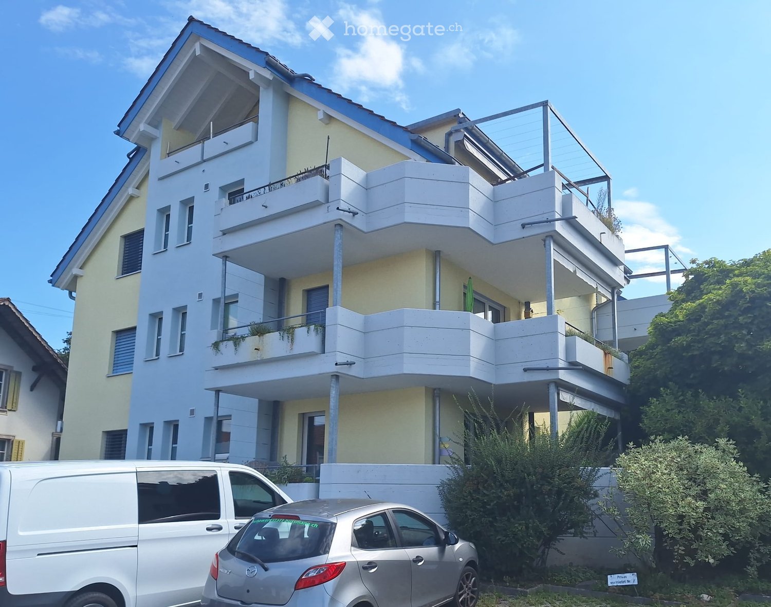 Modern two-story building with white balconies and glass railings, surrounded by greenery and parked vehicles.