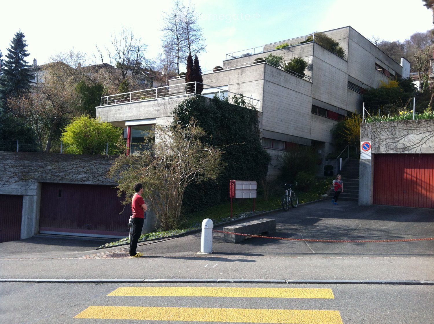 Modern multi-story building with concrete facade, raised ground floor, garage, and surrounding greenery.