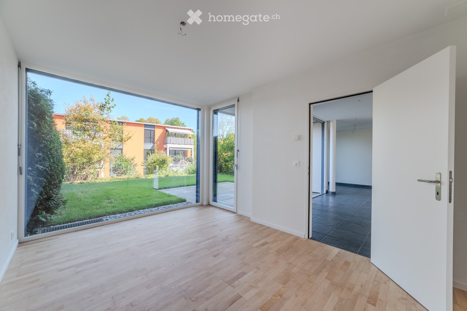 Empty room with light wooden flooring, large glass sliding doors leading to a garden, and a white door to another room.