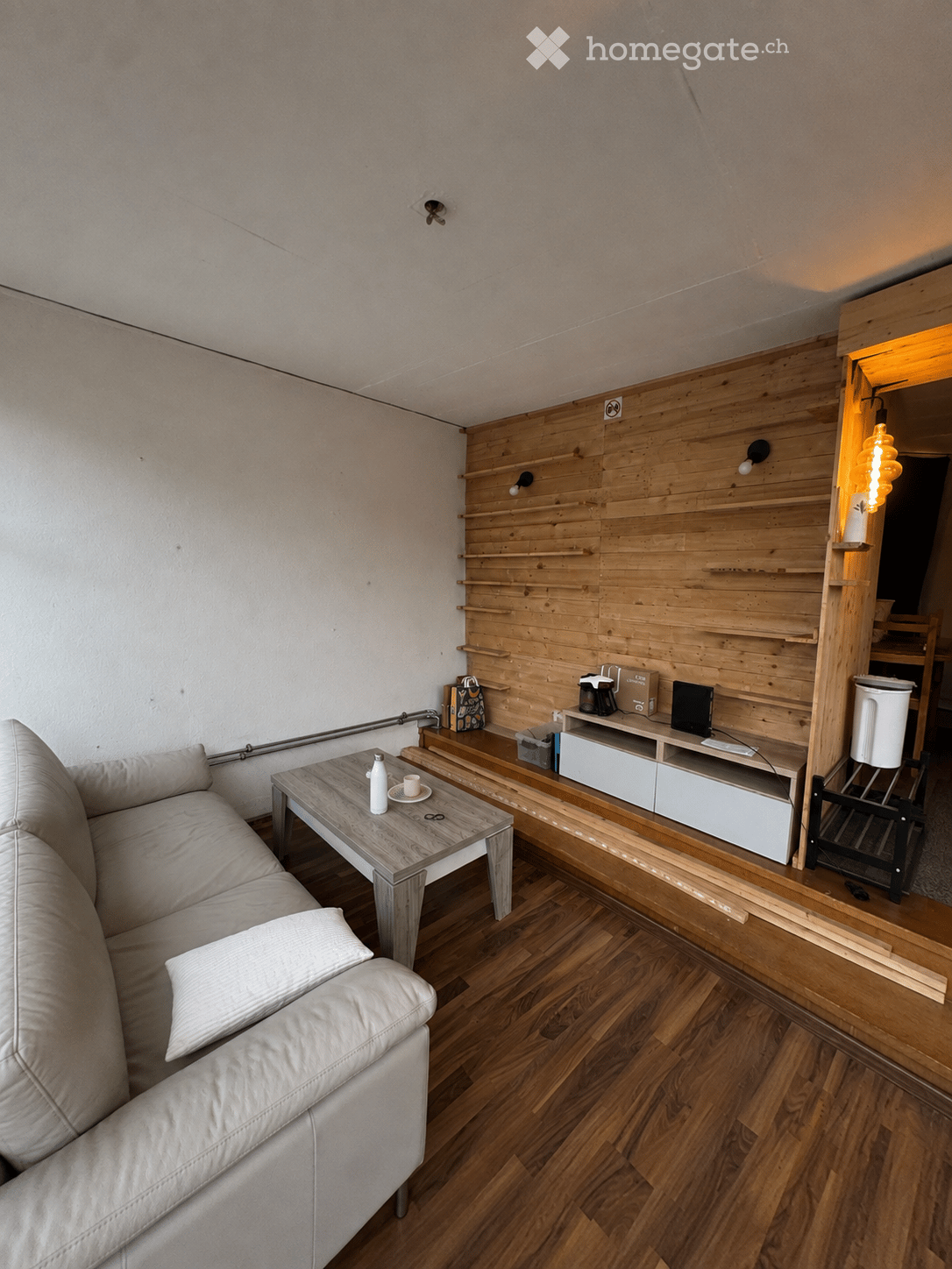 Modern living room with light wooden flooring, white walls, and a wooden accent wall. Features a white leather sofa, a wooden coffee table with a bottle and cup, a TV stand with a laptop, and built-in wooden shelves.
