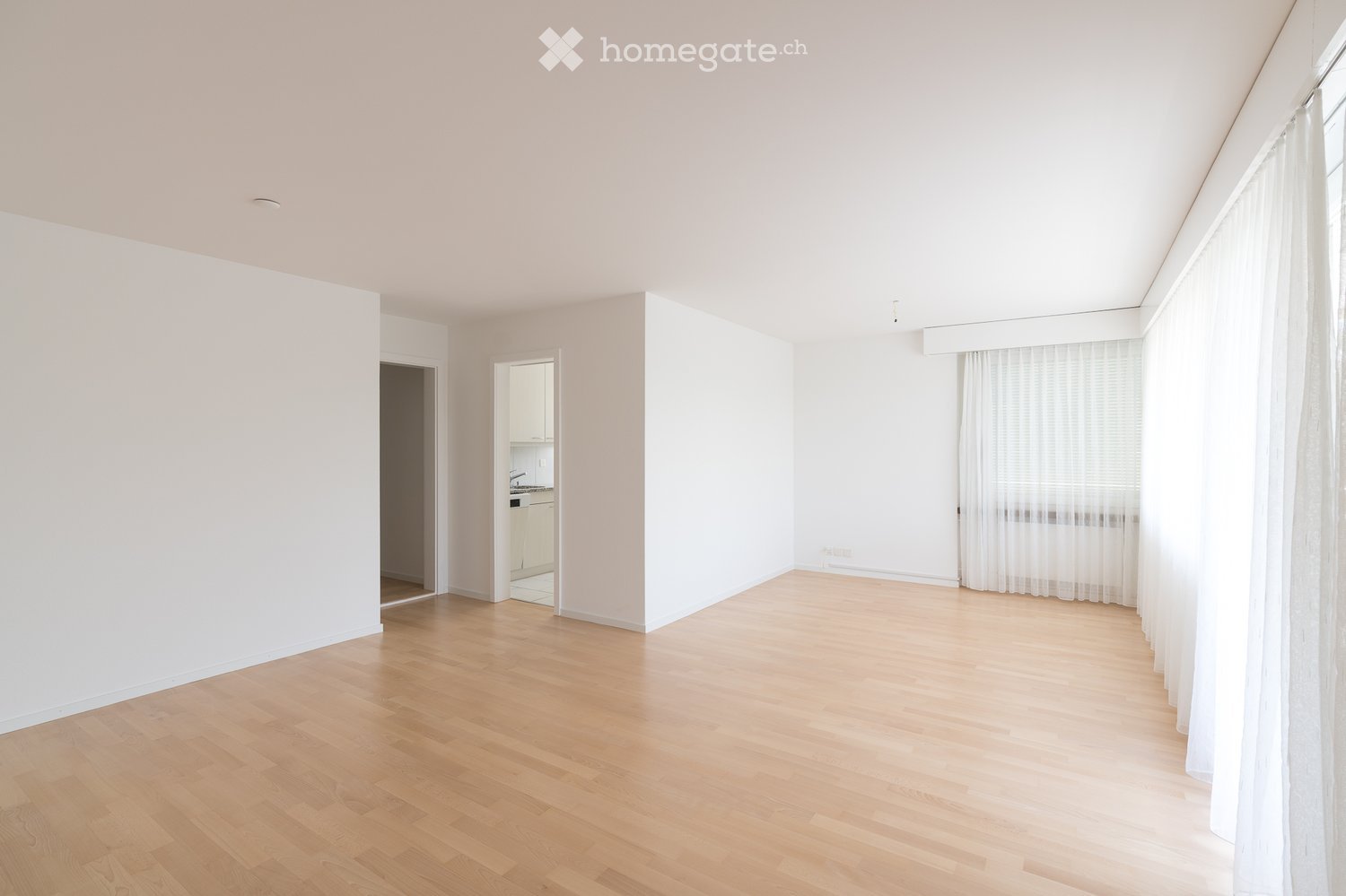 Empty room with light wooden flooring, white walls, and large windows with white curtains. The room is unfurnished and has an open layout with visible doorways leading to other areas.