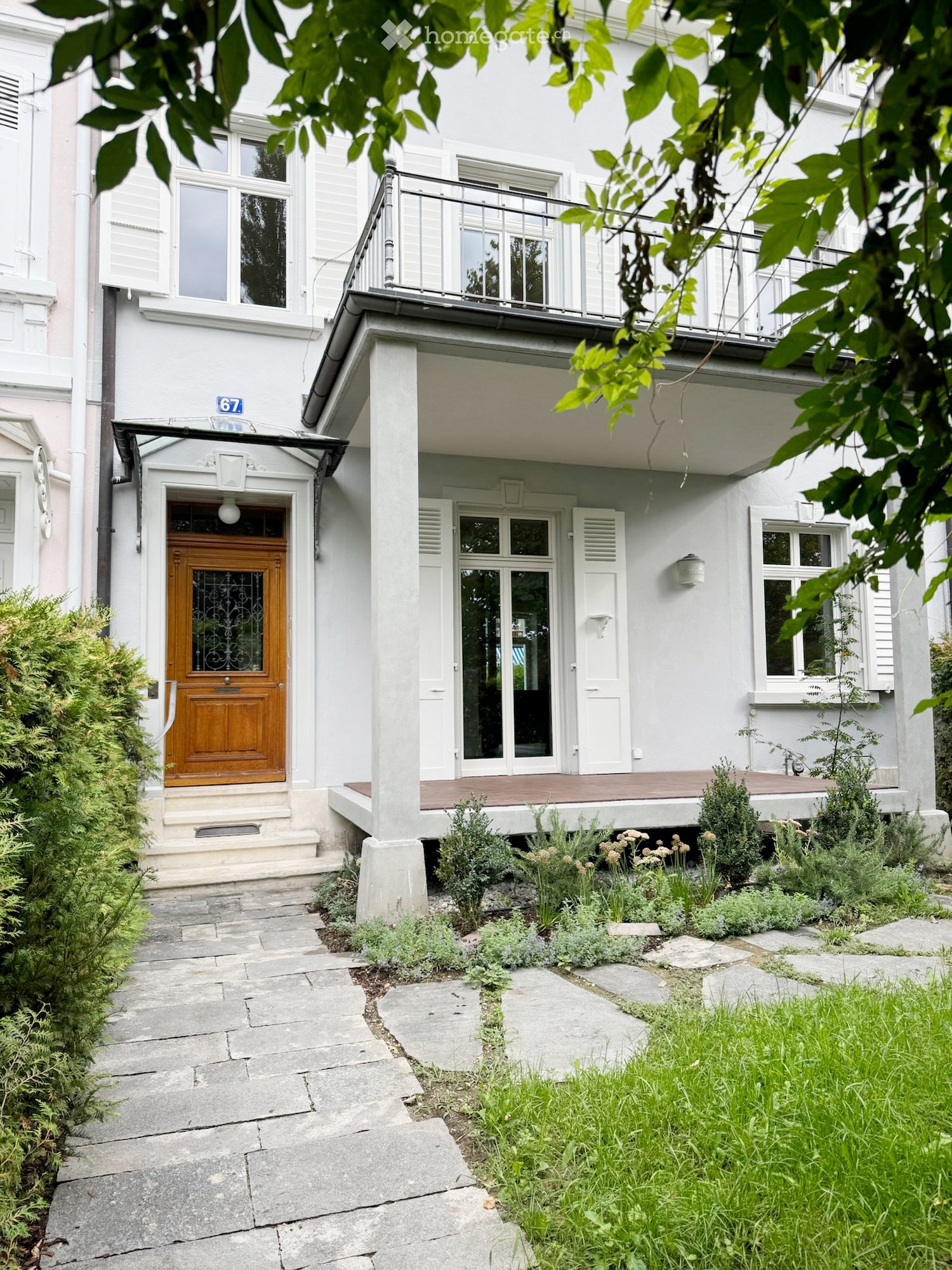 Two-story white building with a wooden front door, balcony, and small garden area with plants and shrubs.