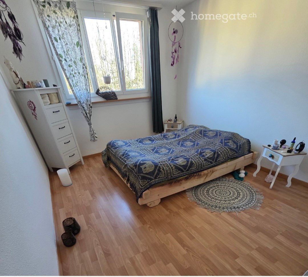 Cozy bedroom with wooden flooring, a large bed with a patterned blue and black bedspread, a nightstand with various items, a white dresser with drawers, and a dreamcatcher hanging on the wall. The room features large windows with sheer curtains and a roun